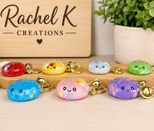 Load image into Gallery viewer, Six cute, rounded, jelly-like keychains in assorted colors (red, yellow, light blue, green, blue, pink, purple) with gold keyrings, displayed on a wooden stand in front of a wooden sign that reads 'Rachel K Creations'.
